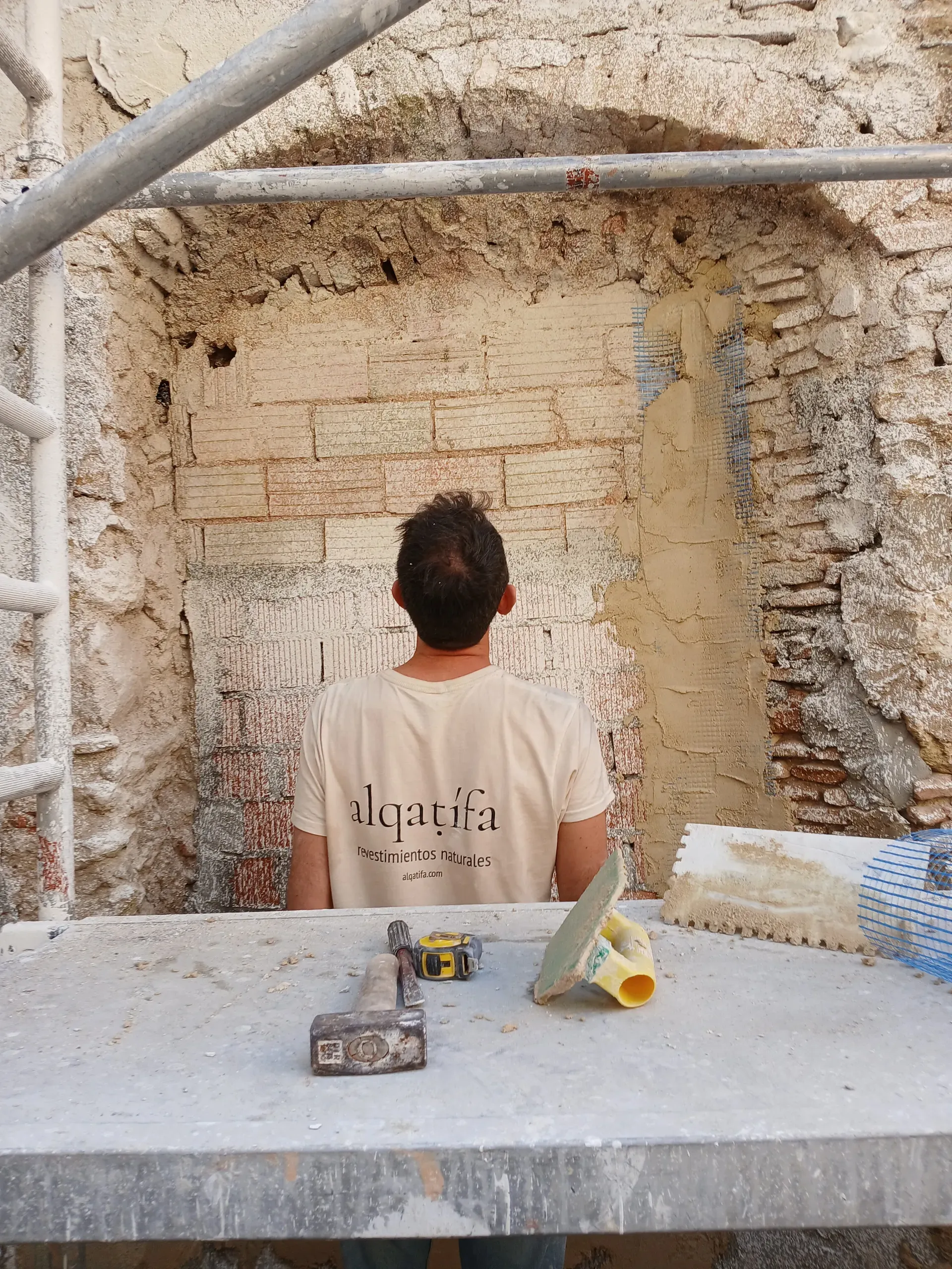Primer plano de herramientas de albañilería sobre mesa de trabajo, con artesano observando el paramento de ladrillo a revocar